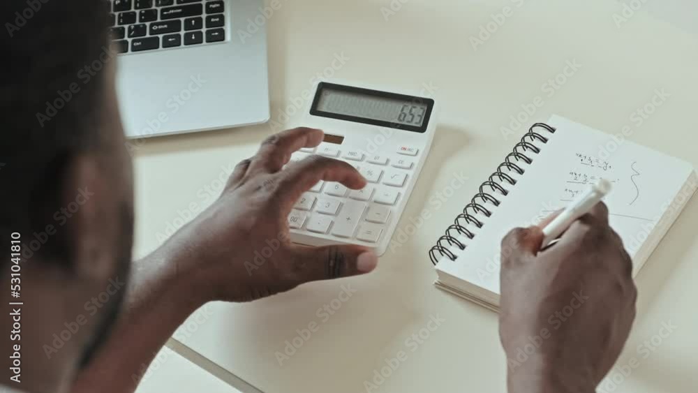 Over the shoulder shot of Black man calculating finances and taking notes in notebook, sitting at desk in home office Over the shoulder shot of Black man calculating finances and taking notes in notebook, sitting at desk in home office