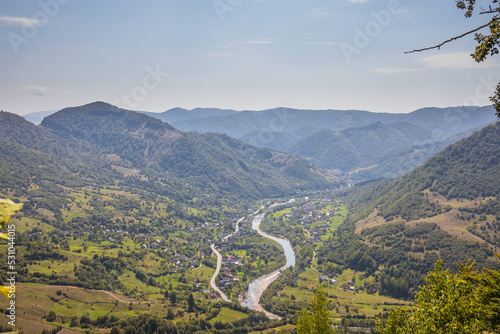 View from a high mountain to a valley between mountains with a river and a village on a summer day. View over the hills, mountains, valley, river and village. Carpathians. Ukraine
