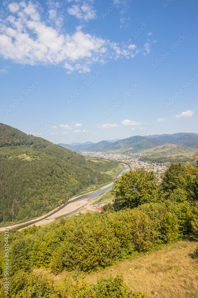 View from a high mountain to a valley between mountains with a river and a village on a summer day. View over the hills, mountains, valley, river and village. Carpathians. Ukraine