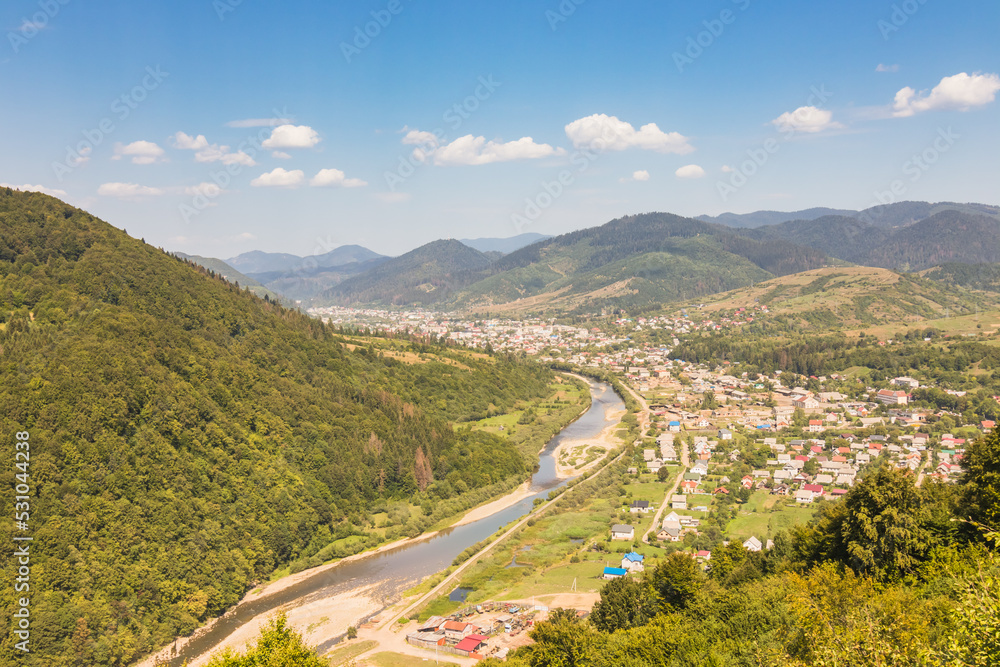 View from a high mountain to a valley between mountains with a river and a village on a summer day. View over the hills, mountains, valley, river and village. Carpathians. Ukraine