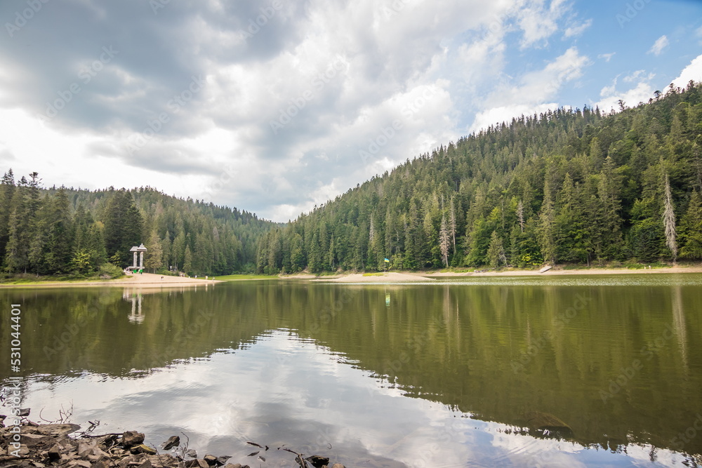 Landscape with calm lake in summer. Forest reflection in the water. beautiful travel background of Synevyr, Ukraine. Tranquil green nature scene. Cloudy outdoor environment