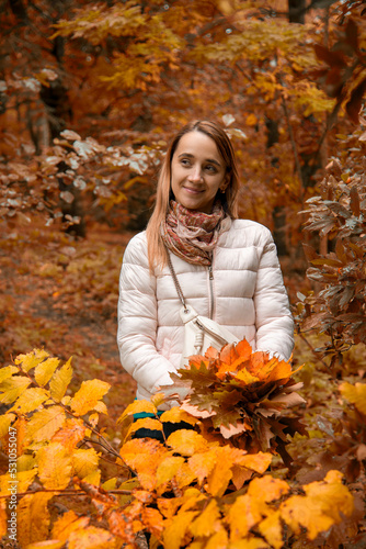 beautiful smiling girl in pink sport jacket walking in autumn park with a bouquet of autumn leaves holding in her hands