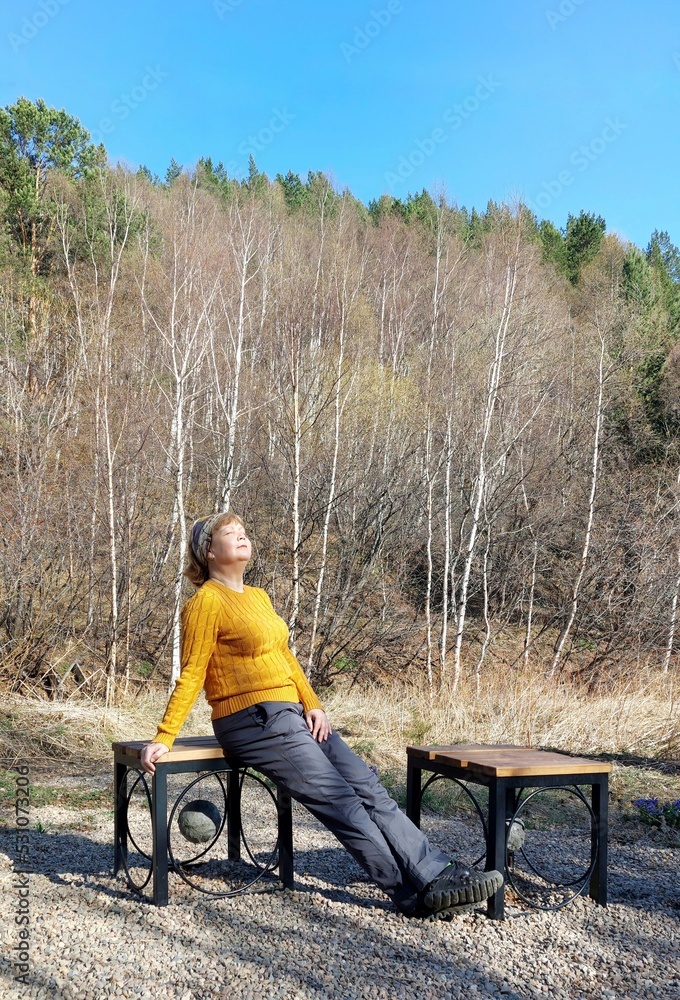 A woman in a yellow jumper is sitting on a bench in a forest park.She turned her face to the sun,enjoying the warmth.Autumn day.