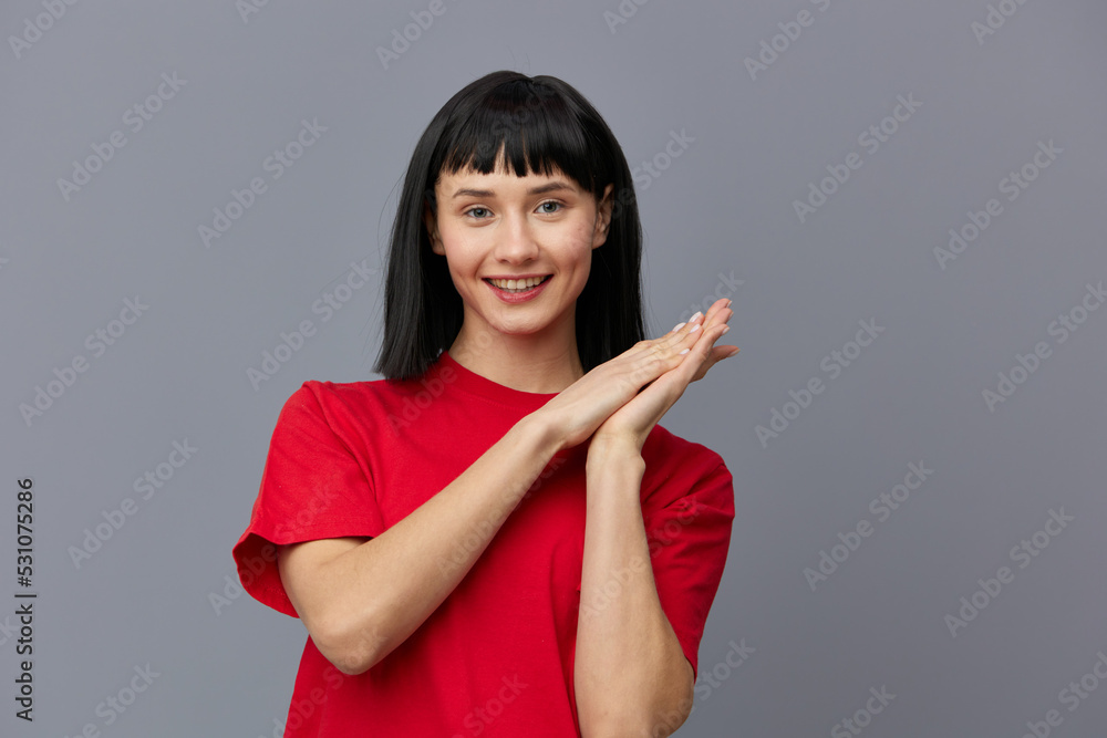 a sweet, modest, attractive woman stands on a gray background in a red T-shirt and smiling pleasantly at the camera keeps her fingers folded in a relaxed pose