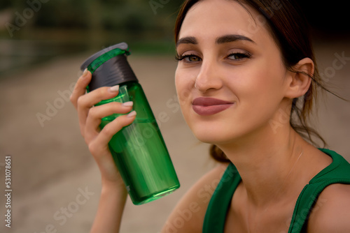 a girl in a sports uniform holds a bottle in her hand, a girl drinks water from a bottle, a girl is dressed in a green tracksuit top and cycling shorts, water balance, outdoor sports, sports lifestyle