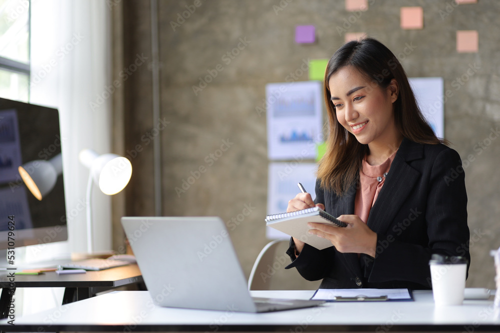 Attractive young Asian business woman in office working on laptop writing work memo on notepad. Attractive young Asian business woman in office working on laptop writing work memo on notepad.