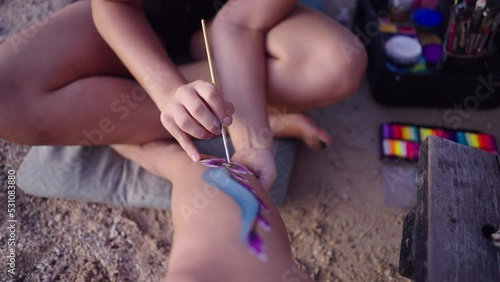 Unrecognizable MUA draws original design with body paint on woman leg at the beach in Koh Phangan, Thailand. 