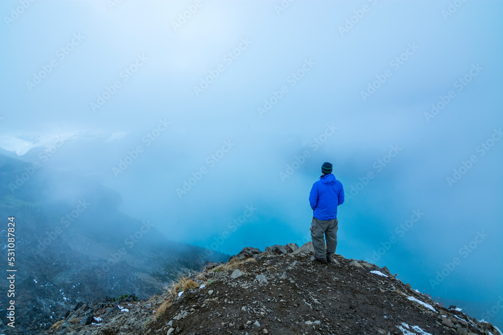 Adventurous athletic male hiker standing at the summit of Panorama Ridge looking down at Garibaldi Lake on a cloudy day.
