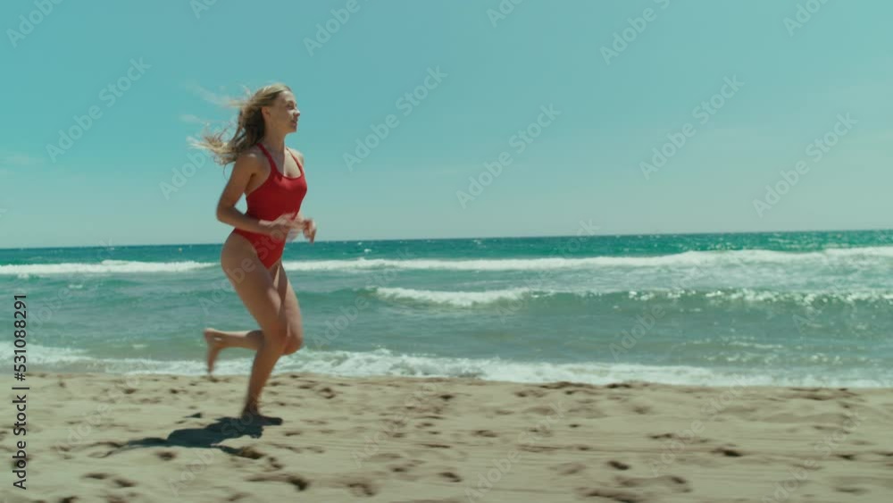 Side tracking shot of fit, athletic and beautiful young woman in red swimsuit run on empty beach in sunshine. Summertime bay coastguard watch concept. Healthy lifestyle of fitness lovers Side tracking shot of fit, athletic and beautiful young woman in red swimsuit run on empty beach in sunshine. Summertime bay coastguard watch concept. Healthy lifestyle of fitness lovers
