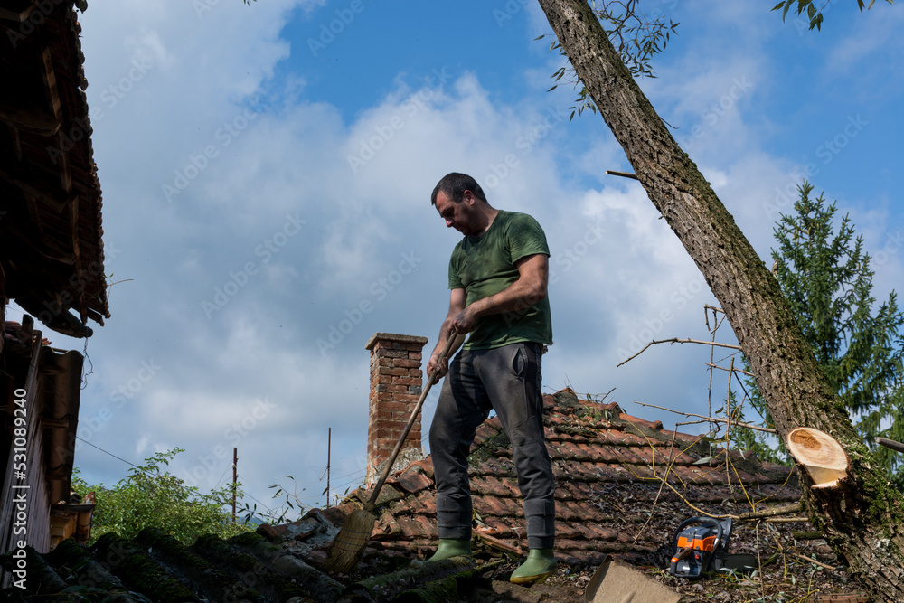 A man stands and cleans with a broom on the roof of the house after a big storm on a summer day. Storm and damage to buildings concept A man stands and cleans with a broom on the roof of the house after a big storm on a summer day. Storm and damage to buildings concept