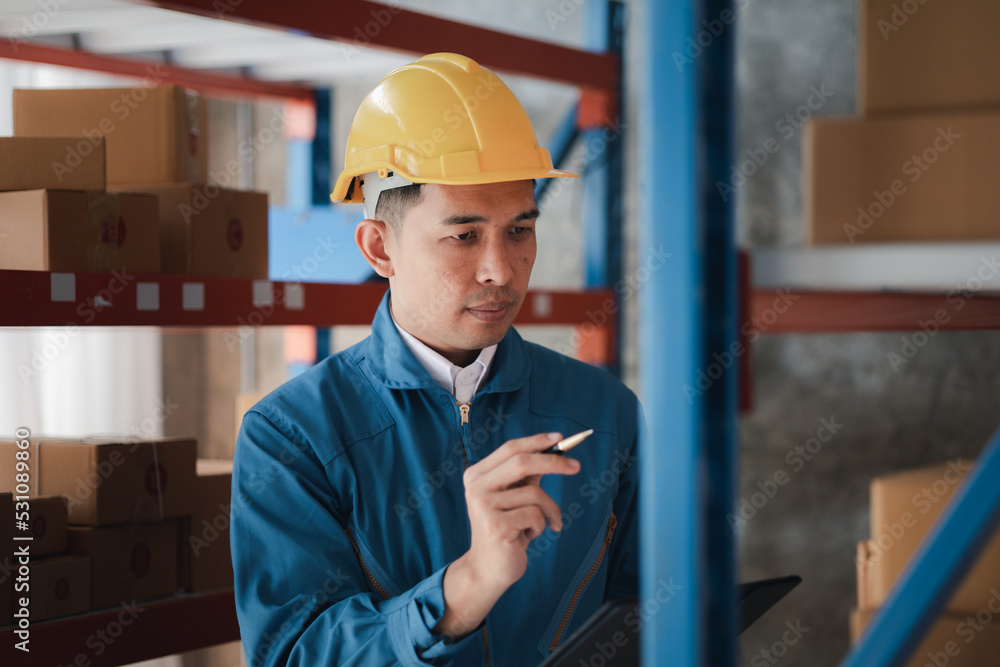 Asian man wearing protective clothing working in a warehouse, he is checking stock, wearing protective clothing while working at a large warehouse according to company policy and worker safety. Asian man wearing protective clothing working in a warehouse, he is checking stock, wearing protective clothing while working at a large warehouse according to company policy and worker safety.
