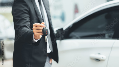 A man in a suit stands holding the car keys next to a white car, getting a new car with a car showroom dealer. Car trading concept. © kamiphotos