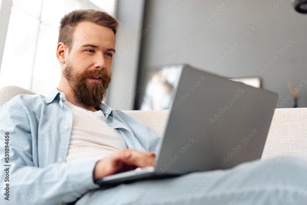 Young bearded man sitting on sofa at home using laptop computer Young bearded man sitting on sofa at home using laptop computer