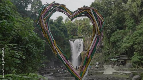 Cute handmade heart made from wooden sticks against huge tropical waterfall on a river in the jungles. Symbol of love in Ubud water cascade in Bali island. Travel Indonesia and explore wild nature.