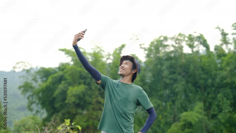 4K, Slow Motion, Young Asian man with a mustache, tall figure wearing his favorite sun hat,He raised his cell phone and took a selfie with the green fields, in front of him, and smiled happily. 4K, Slow Motion, Young Asian man with a mustache, tall figure wearing his favorite sun hat,He raised his cell phone and took a selfie with the green fields, in front of him, and smiled happily.