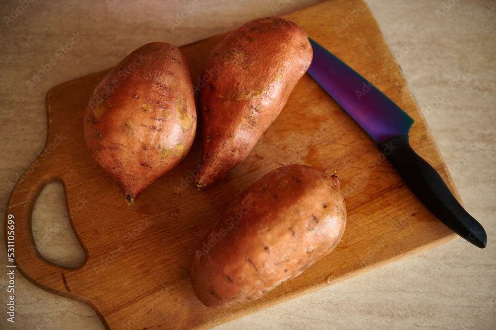 Top view of washed organic sweet potato tubers, batata, yam and kitchen knife on a wooden cutting board on kitchen countertop. Healthy eating. Raw vegan food. Food background. Root vegetable Top view of washed organic sweet potato tubers, batata, yam and kitchen knife on a wooden cutting board on kitchen countertop. Healthy eating. Raw vegan food. Food background. Root vegetable
