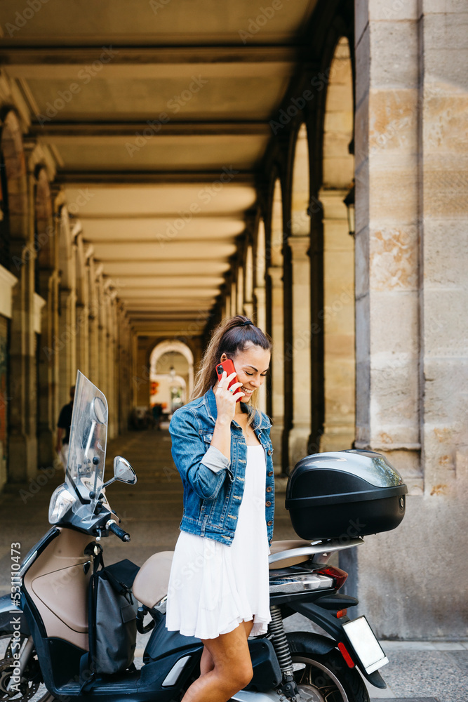Young smiling woman on a motorbike talking on a red smartphone