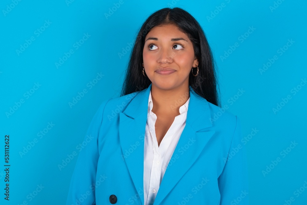 Young latin woman wearing blue blazer blue background looking aside into empty space thoughtful Young latin woman wearing blue blazer blue background looking aside into empty space thoughtful