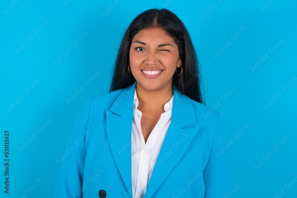 Coquettish Young latin woman wearing blue blazer blue background smiling happily, blinking at camera in a playful manner, flirting with you. Coquettish Young latin woman wearing blue blazer blue background smiling happily, blinking at camera in a playful manner, flirting with you.