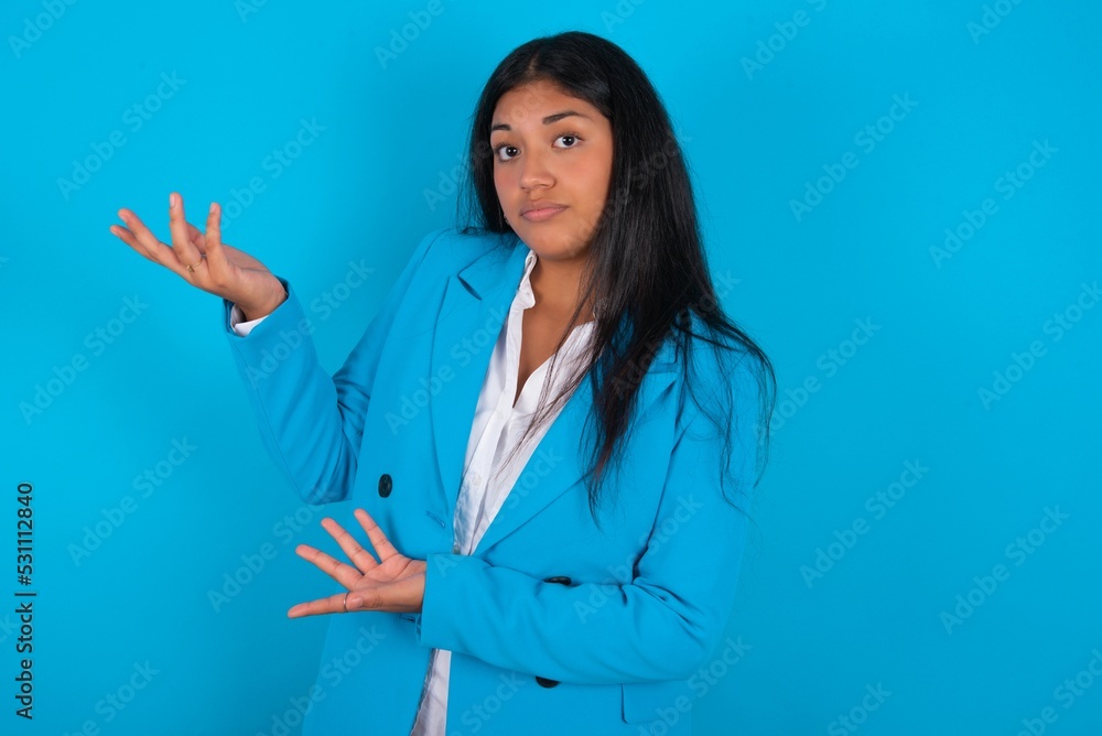 Young latin woman wearing blue blazer blue background pointing aside with both hands showing something strange and saying: I don't know what is this. Advertisement concept. Young latin woman wearing blue blazer blue background pointing aside with both hands showing something strange and saying: I don't know what is this. Advertisement concept.