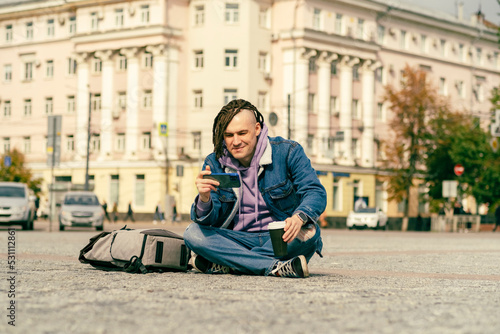 Hipster man with dreadlocks in a denim warm jacket sitting outside on street together with cell phone Outdoors city background. Lifestyle