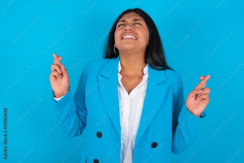 Young latin woman wearing blue blazer blue background gesturing finger crossed smiling with hope and eyes closed. Luck and superstitious concept. Young latin woman wearing blue blazer blue background gesturing finger crossed smiling with hope and eyes closed. Luck and superstitious concept.