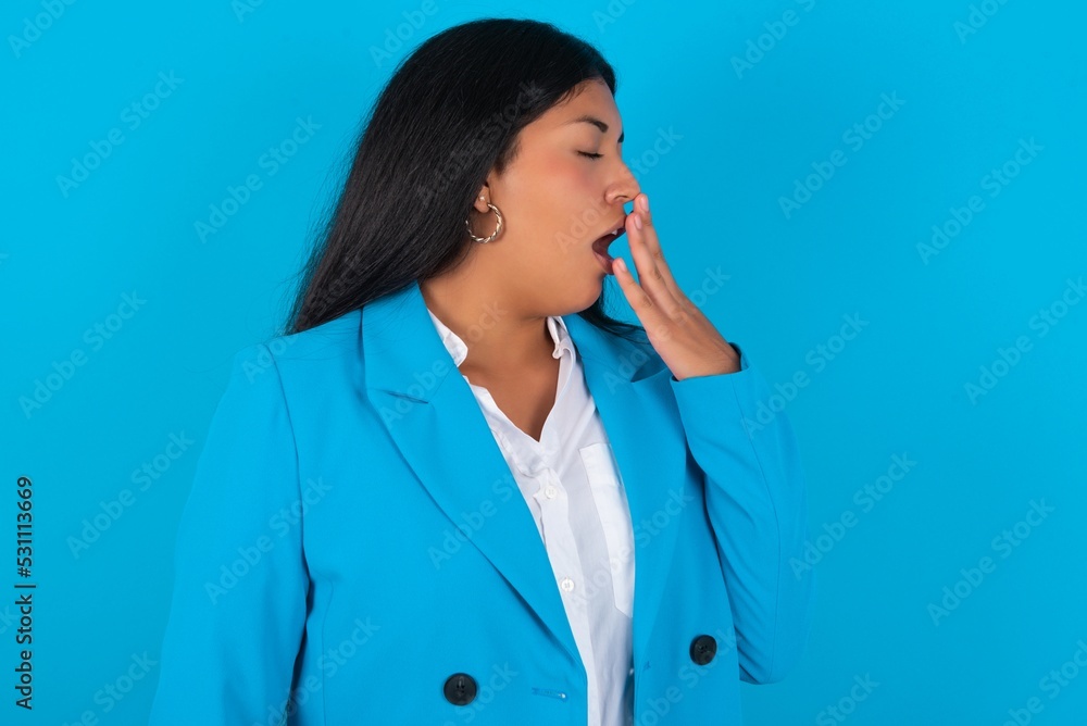Young latin woman wearing blue blazer blue background being tired and yawning after spending all day at work. Young latin woman wearing blue blazer blue background being tired and yawning after spending all day at work.