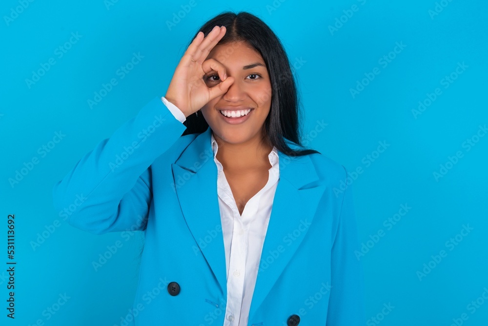 Young latin woman wearing blue blazer blue background doing ok gesture with hand smiling, eye looking through fingers with happy face. Young latin woman wearing blue blazer blue background doing ok gesture with hand smiling, eye looking through fingers with happy face.