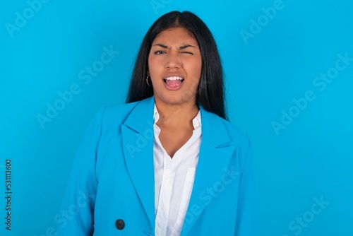 Young latin woman wearing  blue blazer blue background sticking tongue out happy with funny expression. Emotion concept. © Roquillo