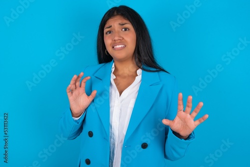 Young latin woman wearing blue blazer blue background afraid and terrified with fear expression stop gesture with hands, shouting in shock. Panic concept.