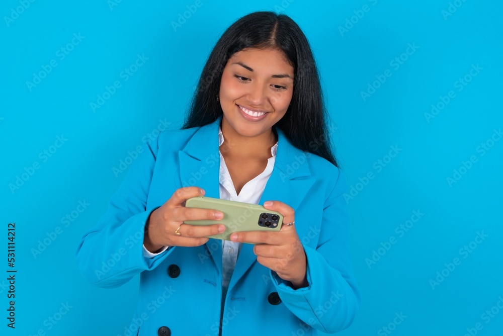 Young latin woman wearing blue blazer blue background holding in hands cell playing video games or chatting Young latin woman wearing blue blazer blue background holding in hands cell playing video games or chatting