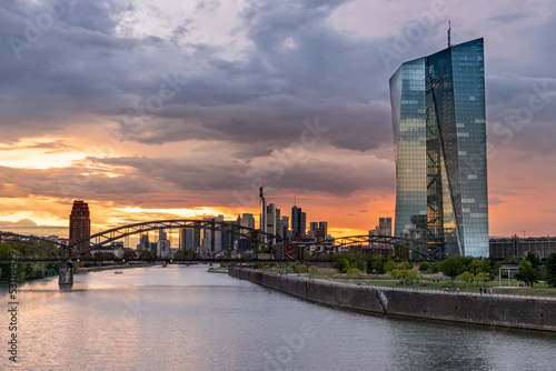 Europäische Zentralbank (EZB) beim Sonnenuntergang mit dunklem Himmel mi Frankfurts Skyline im Hintergrund	