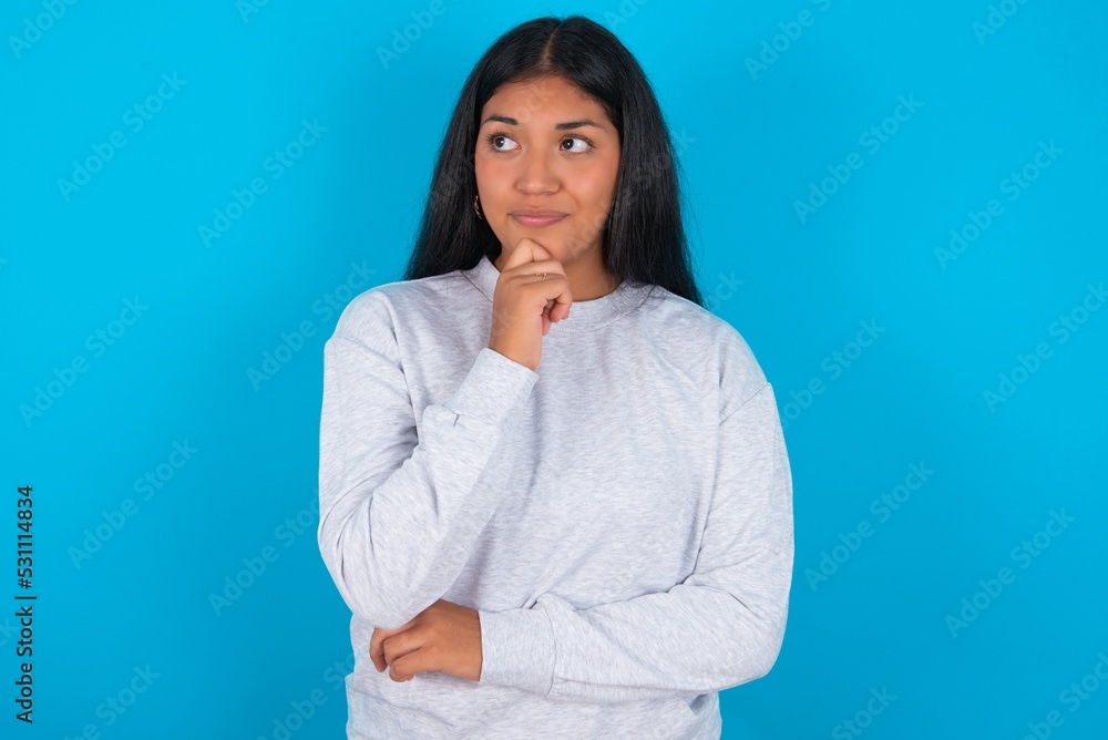Thoughtful Young latin woman wearing gray sweater blue background holds chin and looks away pensively makes up great plan Thoughtful Young latin woman wearing gray sweater blue background holds chin and looks away pensively makes up great plan