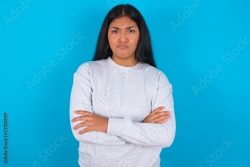Gloomy dissatisfied Young latin woman wearing gray sweater blue background looks with miserable expression at camera from under forehead, makes unhappy grimace