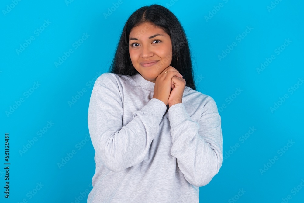 Charming serious Young latin woman wearing gray sweater blue background keeps hands near face smiles tenderly at camera Charming serious Young latin woman wearing gray sweater blue background keeps hands near face smiles tenderly at camera
