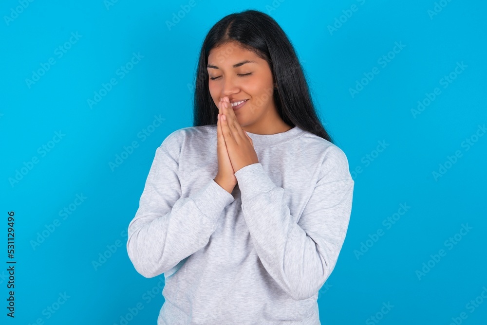 Overjoyed Young latin woman wearing gray sweater blue background laughs joyfully and keeps palms pressed together hears something funny Overjoyed Young latin woman wearing gray sweater blue background laughs joyfully and keeps palms pressed together hears something funny