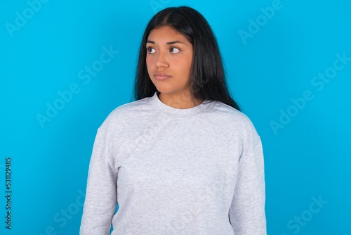 Young latin woman wearing gray sweater blue background stares aside with wondered expression has speechless expression. Embarrassed model looks in surprise