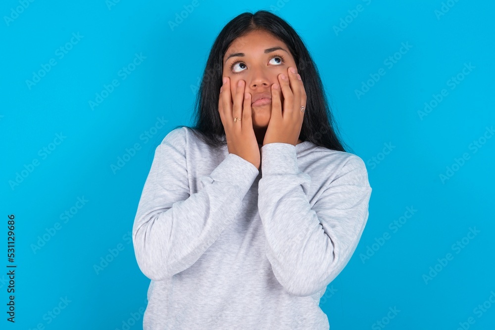 Young latin woman wearing gray sweater blue background keeps hands on cheeks has bored displeased expression. Stressed hopeless model Young latin woman wearing gray sweater blue background keeps hands on cheeks has bored displeased expression. Stressed hopeless model
