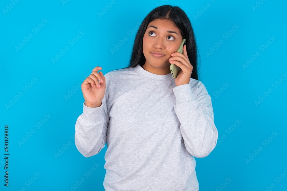 Young latin woman wearing gray sweater blue background speaks on mobile phone spends free time indoors calls to friend. Young latin woman wearing gray sweater blue background speaks on mobile phone spends free time indoors calls to friend.
