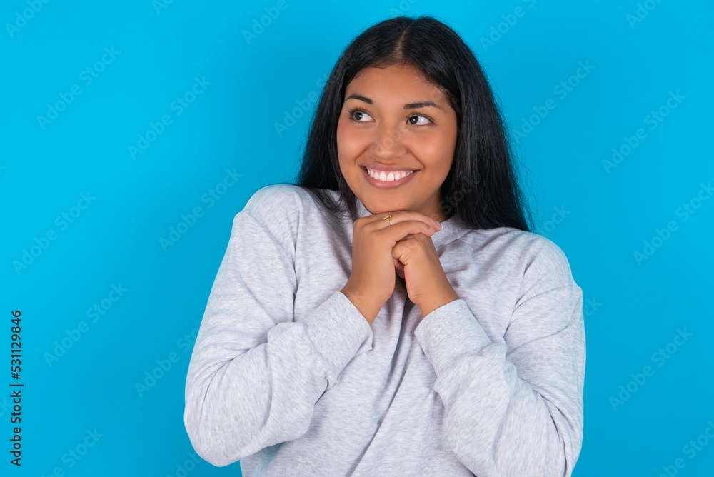 Happy Young latin woman wearing gray sweater blue background anticipates something awesome happen, looks happily aside, keeps hands together near face, has glad expression. Happy Young latin woman wearing gray sweater blue background anticipates something awesome happen, looks happily aside, keeps hands together near face, has glad expression.