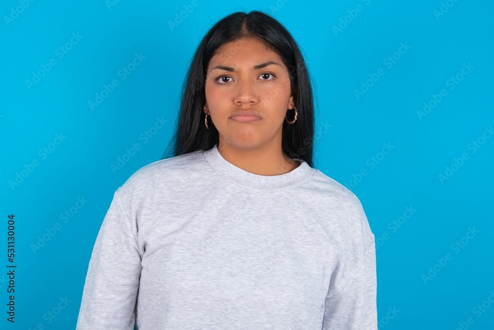 Displeased Young latin woman wearing gray sweater blue background frowns face feels unhappy has some problems. Negative emotions and feelings concept