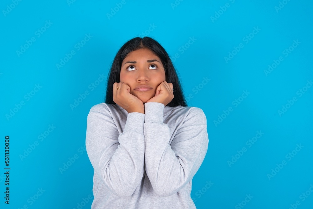 Portrait of sad Young latin woman wearing gray sweater blue background hands face look empty space Portrait of sad Young latin woman wearing gray sweater blue background hands face look empty space