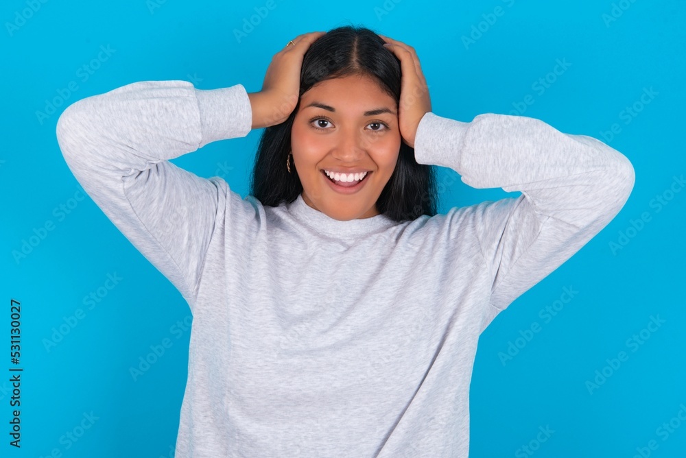 Cheerful overjoyed Young latin woman wearing gray sweater blue background reacts rising hands over head after receiving great news. Cheerful overjoyed Young latin woman wearing gray sweater blue background reacts rising hands over head after receiving great news.