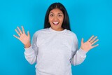 Delighted positive Young latin woman wearing gray sweater blue background opens mouth  and arms palms up after having great result