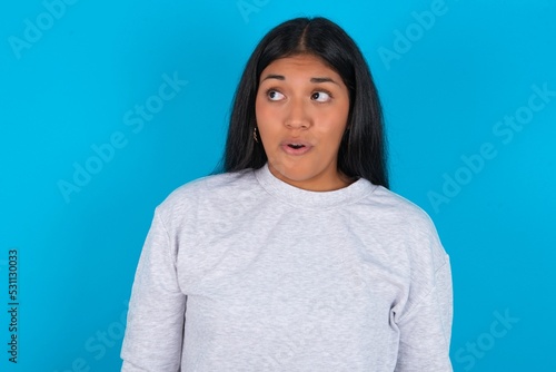 Shocked Young latin woman wearing gray sweater blue background look empty space with open mouth screaming: Oh My God! I can't believe this.