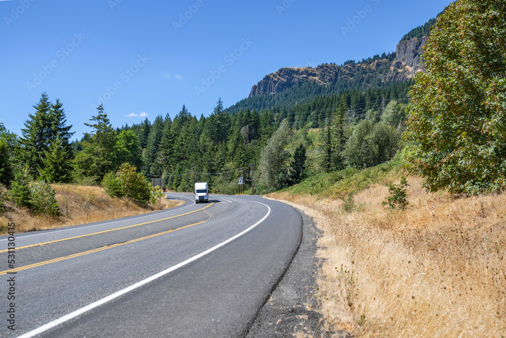 Professional white big rig semi truck tractor with semi trailer running for delivery on the turning highway road in scenic nature wild Columbia Gorge area Professional white big rig semi truck tractor with semi trailer running for delivery on the turning highway road in scenic nature wild Columbia Gorge area