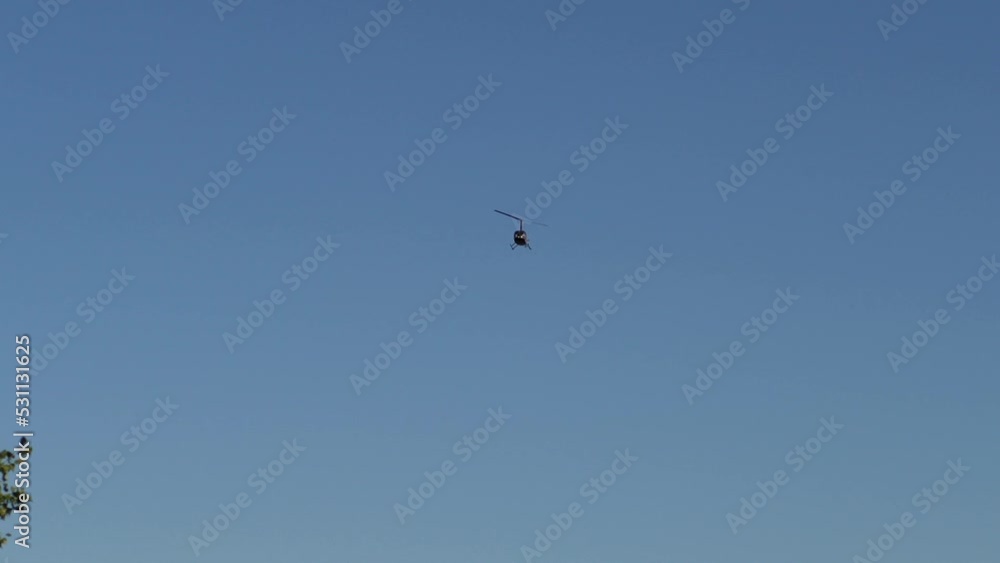 View from ground of civilian helicopter flying across cloudless sky on sunny summer day. Helicopter hiding behind canopy of trees. Shooting in slow motion. View from ground of civilian helicopter flying across cloudless sky on sunny summer day. Helicopter hiding behind canopy of trees. Shooting in slow motion.