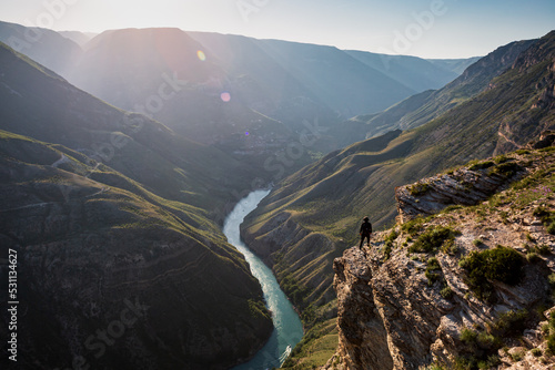 A small figure of a man against the backdrop of high mountains and the Sulak Canyon with a river at sunset.