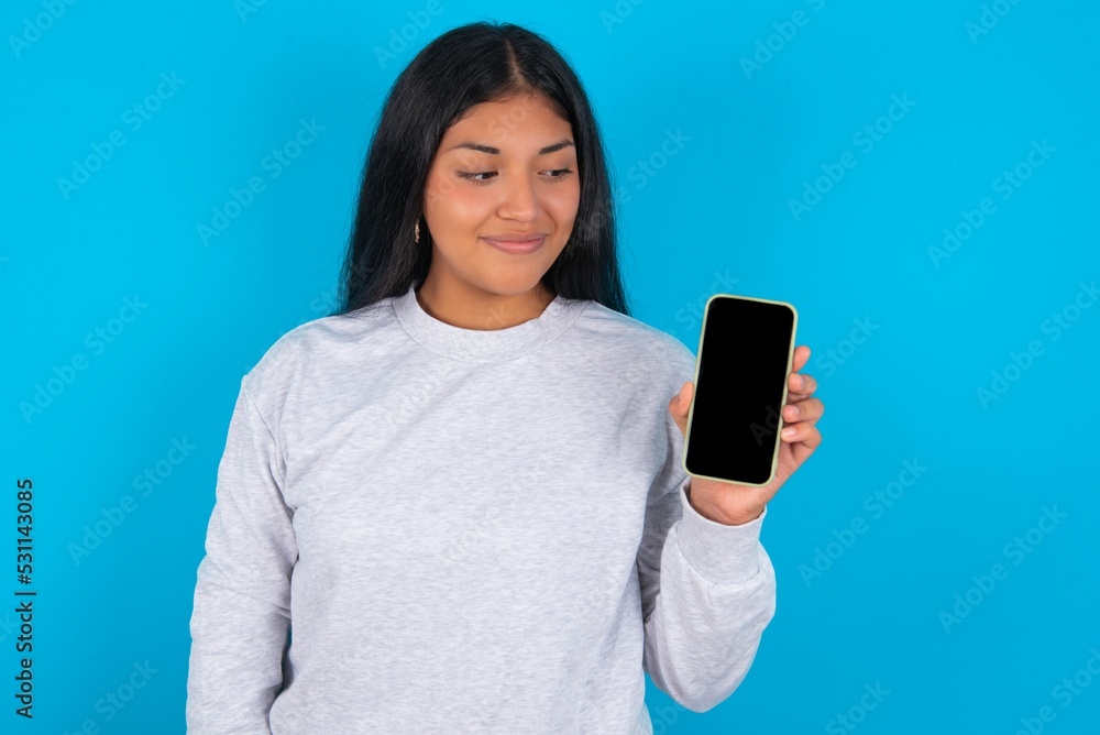 Photo of interested Young hispanic woman wearing grey sweatshirt over blue background hold telephone look side empty space screen Photo of interested Young hispanic woman wearing grey sweatshirt over blue background hold telephone look side empty space screen