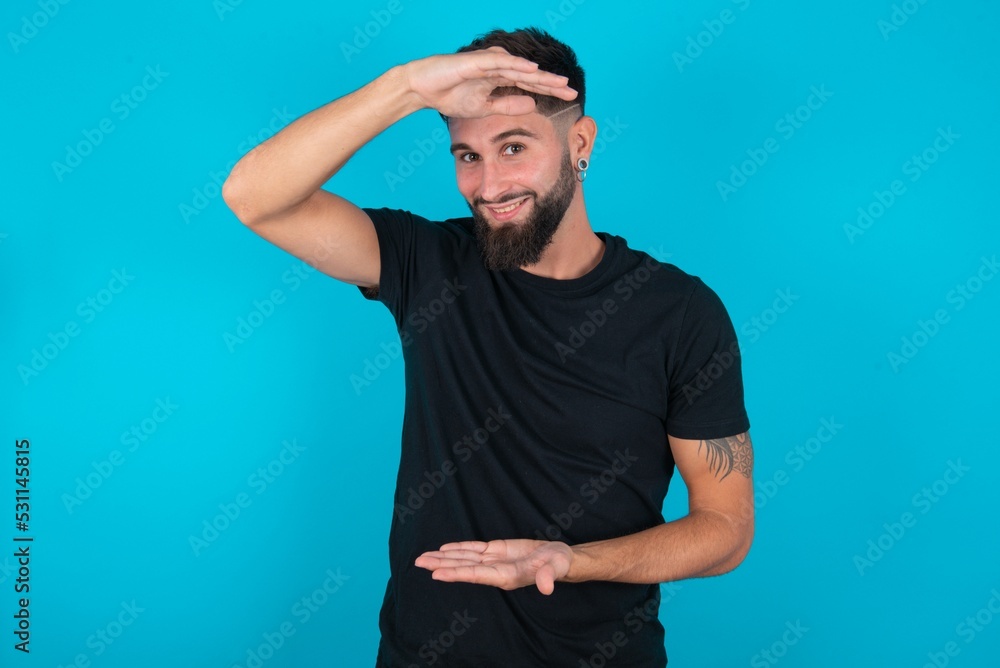 young hispanic bearded man wearing black T-shirt standing against blue background gesturing with hands showing big and large size sign, measure symbol. Smiling looking at the camera. young hispanic bearded man wearing black T-shirt standing against blue background gesturing with hands showing big and large size sign, measure symbol. Smiling looking at the camera.